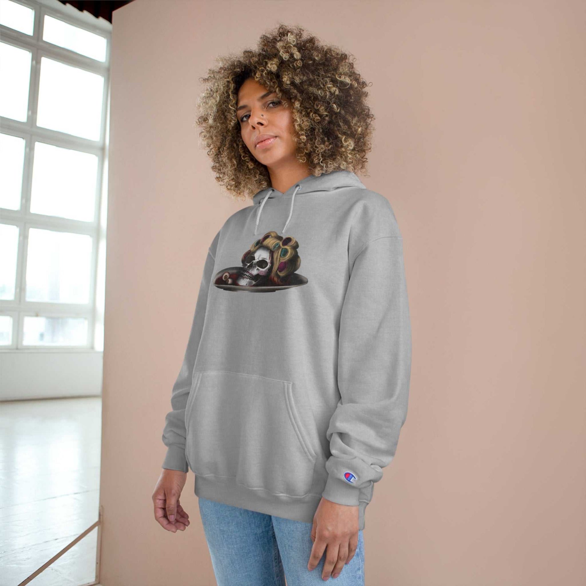Woman wearing a gray Halloween Champion Hoodie with a spooky graphic, leaning against a wall in a bright room.